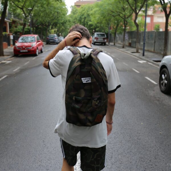 boy wearing white shirt and black shorts carrying backpack standing on black concrete road between vehicles and trees during daytime